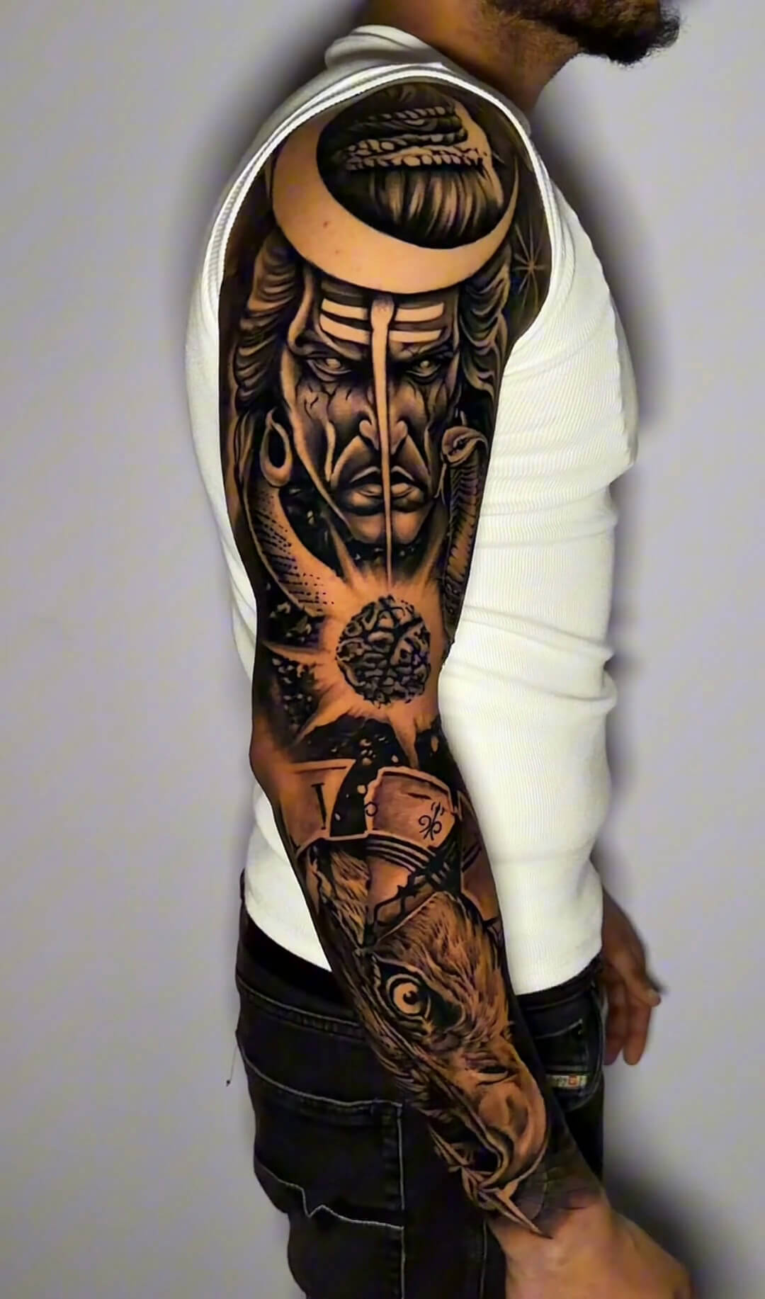 Full-arm tattoo combining a Shiva portrait, crescent, cobra, rudraksha bead and eagle with clock details